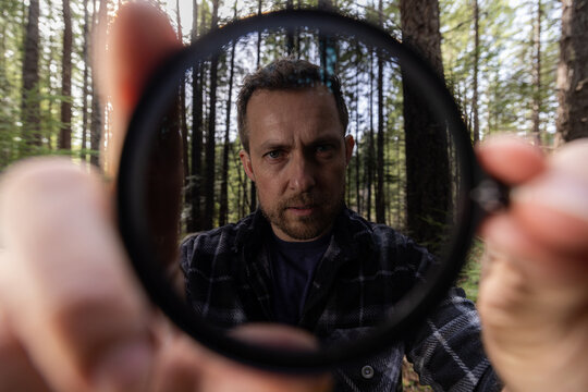 Man smiles through a neutral density filter for the camera . Green trees blur behind his face. Natural light filters through the canopy. He holds the camera filter near the lens.