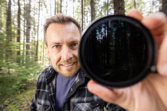 Man smiles through a neutral density filter for the camera . Green trees blur behind his face. Natural light filters through the canopy. He holds the camera filter near the lens.