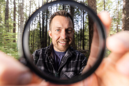 Man smiles through a neutral density filter for the camera . Green trees blur behind his face. Natural light filters through the canopy. He holds the camera filter near the lens.