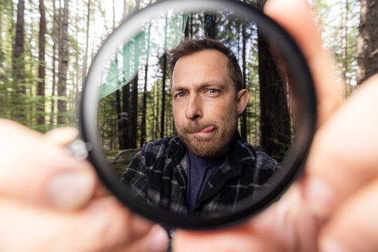 Man smiles through a neutral density filter for the camera . Green trees blur behind his face. Natural light filters through the canopy. He holds the camera filter near the lens.