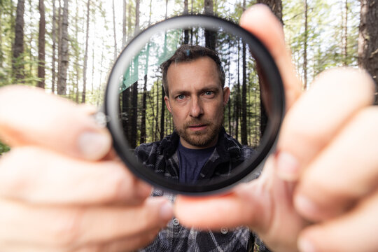 Man smiles through a neutral density filter for the camera . Green trees blur behind his face. Natural light filters through the canopy. He holds the camera filter near the lens.