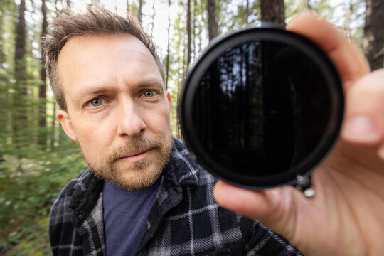 Man smiles through a neutral density filter for the camera . Green trees blur behind his face. Natural light filters through the canopy. He holds the camera filter near the lens.