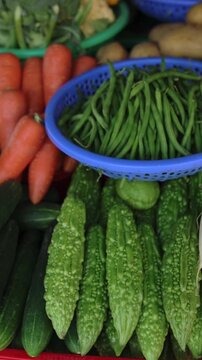 Vertical screen Vibrant Vietnam Street Market Vegetables Stall, Colorful Closeup Of Bitter Gourd Rows, Long Beans In Blue Basket, Cucumbers, Carrots, White Radishes And Potatoes, Dewy Skin Texture,