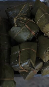 Vertical screen Vietnam Street Market Display Banana Leaf Wrapped Rice Cakes Stacked On Metal Tray Tied With Natural String Closeup Of Glossy Green Leaves, Textured Folds, Rustic Market Lighting,