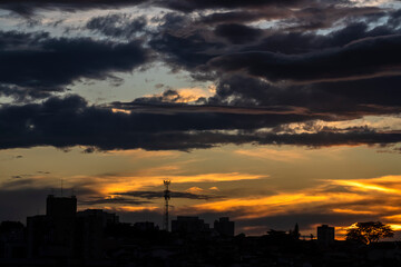 A dramatic golden sunset casts a vibrant glow over a city skyline. Dark clouds part to reveal the brilliant sun, silhouetting buildings and a lone tree against the luminous sky.