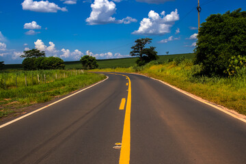 Naklejka premium Asphalt road with yellow lines curves through the vibrant green landscape of rural Brazil under a blue sky. A concept of journey, travel, and perspective in South America's countryside.