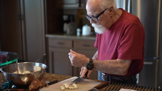 Senior man mashes garlic in his kitchen as he prepares a dish using fresh ingredients and a sharp knife.