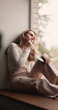 Pensive young adult woman sitting on windowsill alone at home, holding smartphone, waiting for call, feeling sadness, loneliness or boredom, thinking on personal issues during weekend leisure time