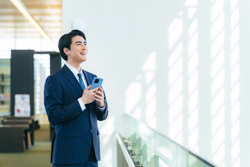 Fototapeta premium オフィスビルのロビーでスマホを持って遠くを眺める男性ビジネスパーソン