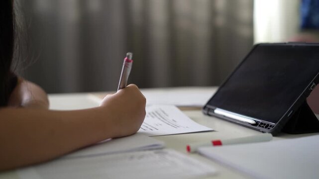 closeup hand of child learn from home write on paper or young kid girl student writing doing homework with computer tablet on desk by study online class or people back to school or business learning