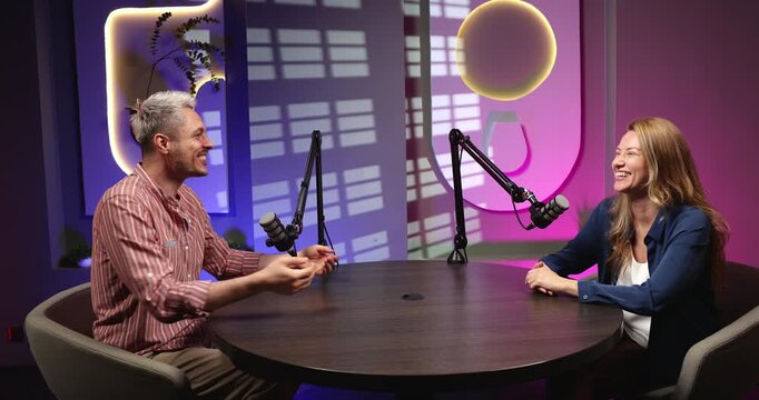 Man and woman sit at table with microphones on stands, talking, smiling during podcast recording in modern studio with neon lighting. Media creation, communication, storytelling, digital broadcasting