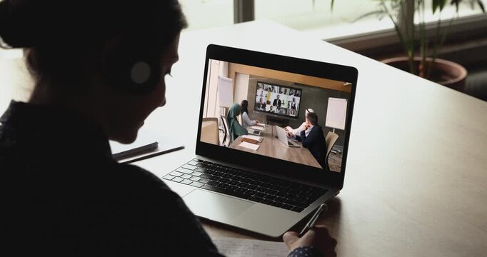 Business professional woman attending international hybrid business conference, talking notes, looking at laptop screen with Arabian colleagues sitting at online group video call board in meeting room