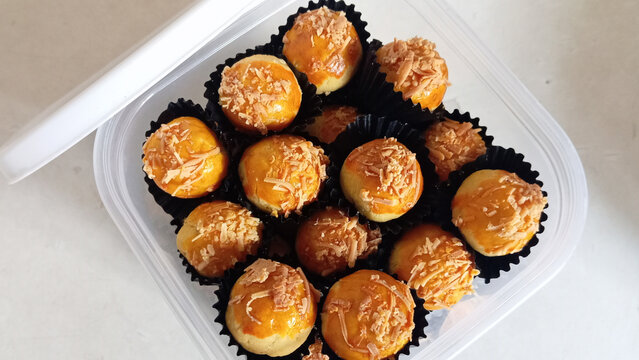 Close-up of homemade mini cheese ball pastries topped with shredded cheese, arranged in black paper cups inside a transparent plastic container on a white table. A popular sweet snack often served for