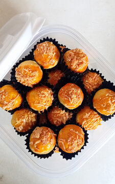 Close-up of homemade mini cheese ball pastries topped with shredded cheese, arranged in black paper cups inside a transparent plastic container on a white table. A popular sweet snack often served for