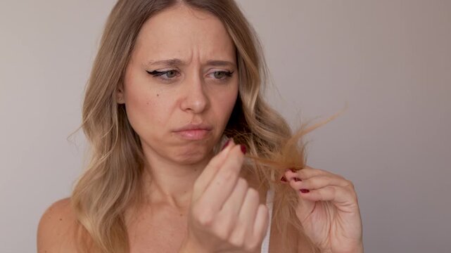 Close-up of young blonde woman holding and examining damaged hair ends with worried expression. Split ends, dryness and hair damage concept.