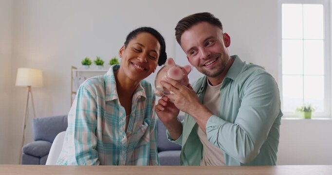 Smiling multiethnic couple sitting at table at home playfully holding piggy bank shaking it, enjoy ringing of coins as symbol of savings and enough of money. Achieving financial independence