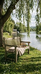 A serene wooden bench by a river features a stack of books and a blanket under the shade of a weeping willow tree.