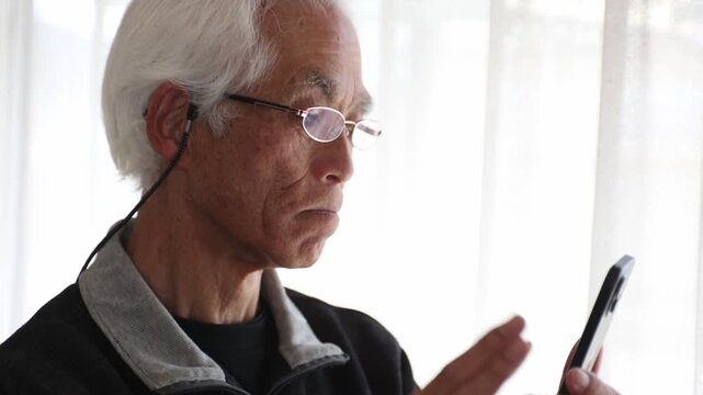 Senior Japanese man with glasses using smartphone at home, focused expression, lifestyle, silver generation, farsightedness, elderly, age 60s-70s, side view, natural light.