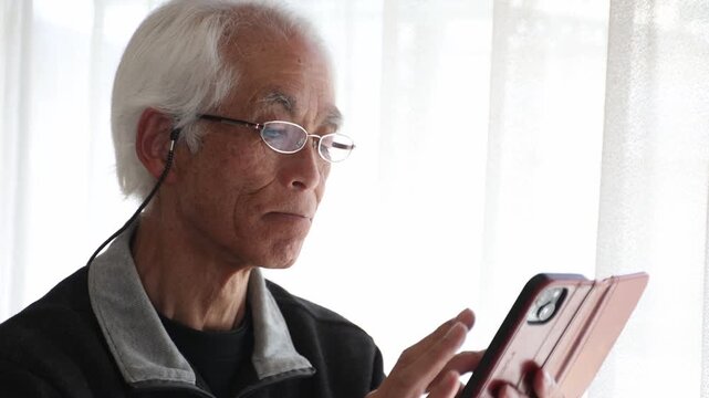 Senior Japanese man with glasses using smartphone at home, focused expression, lifestyle, silver generation, farsightedness, elderly, age 60s-70s, side view, natural light.