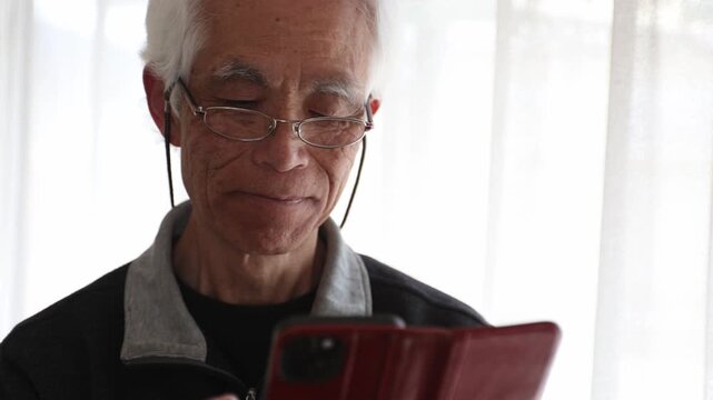 Senior Japanese man with glasses using smartphone at home, focused expression, lifestyle, silver generation, farsightedness, elderly, age 60s-70s, side view, natural light.