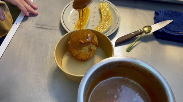Close-up of fried ice cream ball topped with warm caramel sauce during dessert preparation, showing crispy coating, sweet topping and banana in a kitchen setting