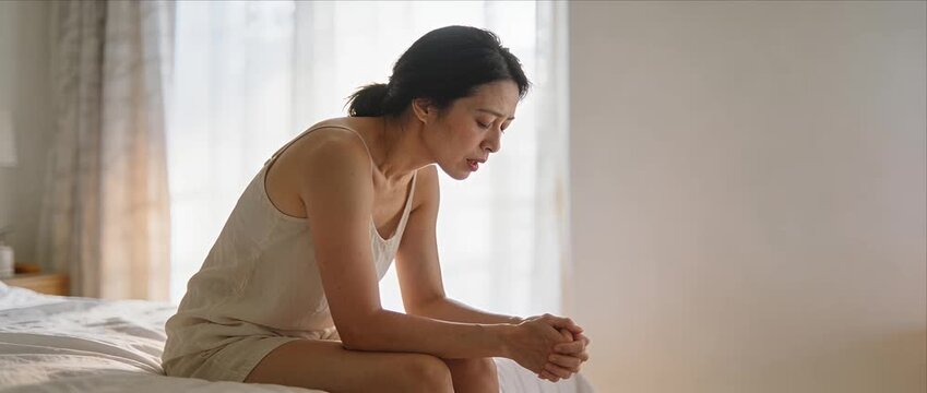 Asian woman middle age sad stress worry depression anxiety sitting on bed in bedroom morning light, insomnia fatigue burnout mental health loneliness despair solitude indoors at home
