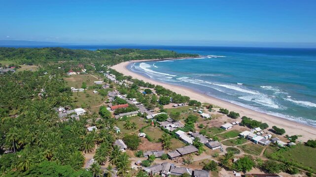 Aerial drone footage of Sawarna surfing spot beach, in Banten, Java island, Indonesia, with many houses and touristic accommodations in the front, blue water and waves, sunny weather