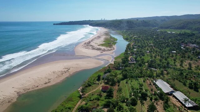 Aerial drone footage of Sawarna surfing spot beach, in Banten, Java island, Indonesia, with many houses and touristic accommodations in the front, blue water and waves, river behind the sand