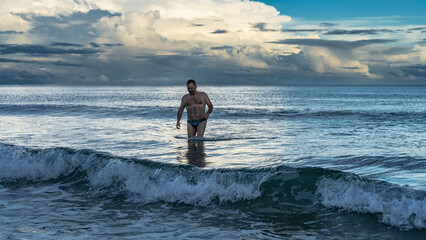 Early morning. Dawn over the ocean. A man in swimming trunks stands in the foaming waves. Reflection. The clouds in the blue sky are highlighted in gold. Malaysia. Borneo. Kota Kinabalu. © Вера 