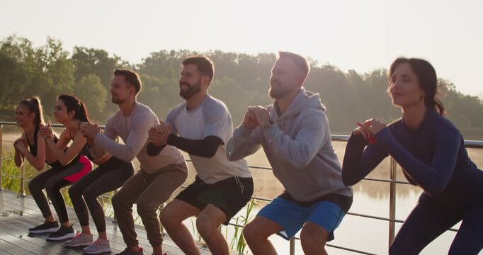 Group of healthy people having an outdoor fitness workout together. Happy smiling male and female athletes doing squats while standing on the bridge together. Sports exercise concept
