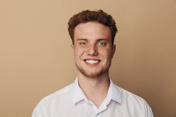Fototapeta premium Smiling young man with curly red hair and freckles wears white shirt against beige background. Portrait of confident male model with natural skin and friendly expression in studio lighting.