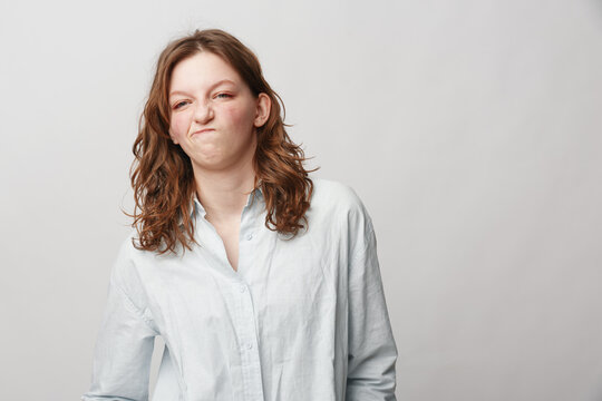 Young woman with curly hair wearing white shirt makes funny face with scrunched nose and pursed lips. Isolated on light background. Concept of humor, expression, and casual style.