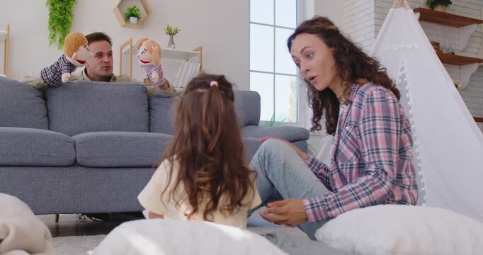 Happy playful family at home play puppet theater show with child girl. Parents and daughter animate plush characters on sofa, laughing together indoors. Family bonding and leisure concept.