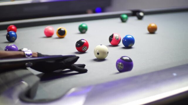 Vibrant close-up on a pool table showing colorful billiard balls. A player's gloved hand and cue stick are poised for a strategic shot, capturing focused gameplay and recreation