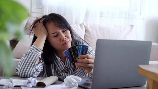 Stressed Asian woman holding multiple credit cards while looking at her laptop, feeling overwhelmed by debt, financial problems, and high expenses at home. Concept of overspending and bankruptcy.