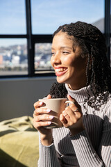 African American woman in ribbed turtleneck holding ribbed mug, smiling, looking out paned window