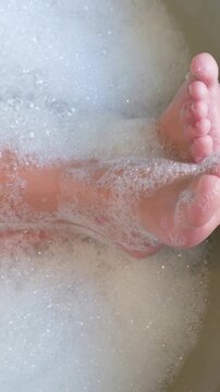 Vertical video: Reacting to water, small child feet wiggling in bathtub, bubbles clinging