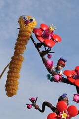 Fototapeta premium LEGO Friends figure of Mia hanging on LEGO Botanicals Plum Blossom branch, handmade knitted funny yellow caterpillar hanged on branch tip. Blue skies in background, sunlit by spring morning sunshine.