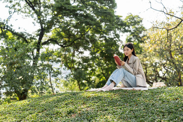 Fototapeta premium Young asian woman relaxing reading book sitting at public park life style outdoor. Happy female Take a break enjoy time living in the nature. Human and nature together