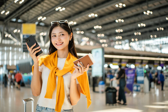 Happy young asian woman traveler holding boarding pass ticket, passport and smartphone at the airport terminal with her luggage and smiling at camera, cheerful tourist female having holiday trip.