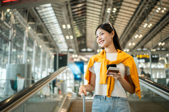 Happy young asian woman traveler holding boarding pass ticket, passport and smartphone at the airport terminal with her luggage and smiling at camera, cheerful tourist female having holiday trip.