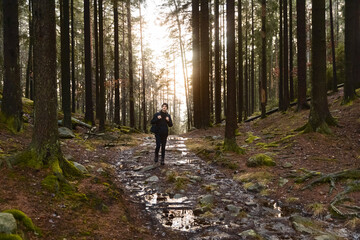 Fototapeta premium Young man in black outdoor gear and beanie walking on a muddy, rocky path through a dense pine forest at sunset. Golden sunlight filters through tall trees, creating a moody, atmospheric woodland.