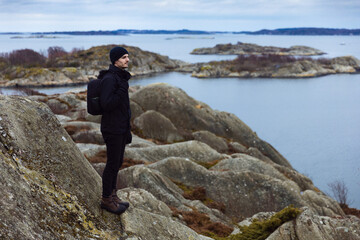 Obraz premium A man in black outdoor clothing with a backpack stands on a rocky cliff, looking out over a calm sea and scenic archipelago islands under a cloudy sky. Perfect for travel and nature themes.