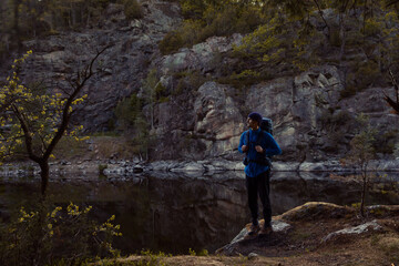 Obraz premium A hiker with a backpack and beanie stands by a calm mountain lake, reflecting the rugged cliffside at dusk. A serene moment of outdoor adventure, trekking, and exploration in the wild wilderness.