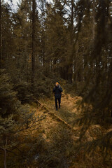 Obraz premium A lone male hiker with a backpack and orange beanie walks through a dense, moody evergreen forest during autumn, surrounded by tall pine trees, mossy ground, and a quiet, wilderness atmosphere.