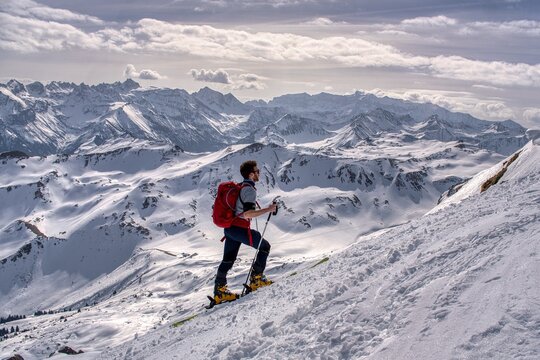 Skier Traversing Vast Snowy Alpine Slope, Lone Backcountry Guide Skiing Across Windswept Plateau Toward Jagged Peaks, Crisp Blue Sky, Sharp Shadows, Visible Tracks, Highaltitude.