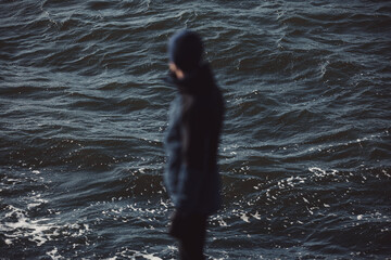 Cinematic view of a blurred silhouette of a man in a beanie looking out over dark, moody, and choppy sea water. Abstract atmospheric photography exploring themes of loneliness, nature, and mystery. © Francis