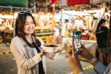 Asian woman enjoy eating noodles street food at night market. Traveler Asian blogger women Happy...