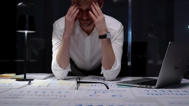 Stressed businessman suffering from overwhelming burnout and work overload, working late into the night, feeling frustrated with deadline pressure in a dark office environment
