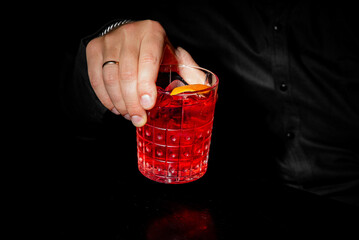 A man's hand with a wedding ring holds a crystal glass of vibrant red Negroni cocktail with an...